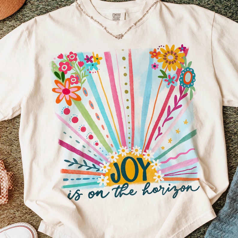 May include: A white t-shirt with a colorful graphic design of a sunrise with flowers and the text "JOY is on the horizon".