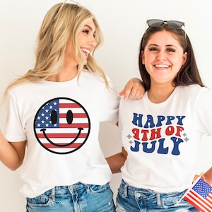 K&ouml;nnte beinhalten: Zwei Frauen tragen wei&szlig;e T-Shirts mit Designs zum 4. Juli. Ein Shirt zeigt ein Smiley mit dem Design der amerikanischen Flagge. Das andere Shirt sagt "Happy 4th of July" in einer Retro-Schrift. Beide Frauen l&auml;cheln und halten amerikanische Flaggen.