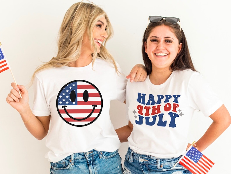 K&ouml;nnte beinhalten: Zwei Frauen tragen wei&szlig;e T-Shirts mit Designs zum 4. Juli. Ein Shirt zeigt ein Smiley mit dem Design der amerikanischen Flagge. Das andere Shirt sagt "Happy 4th of July" in einer Retro-Schrift. Beide Frauen l&auml;cheln und halten amerikanische Flaggen.