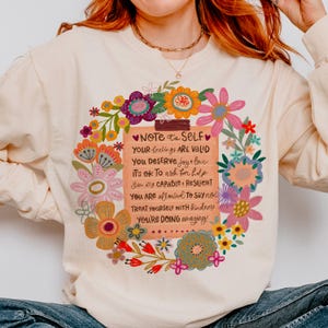 May include: Cream-colored sweatshirt with a floral design and the text "NOTE to SELF." The text includes affirmations such as "Your feelings are valid" and "You deserve joy & love." The design features colorful flowers.