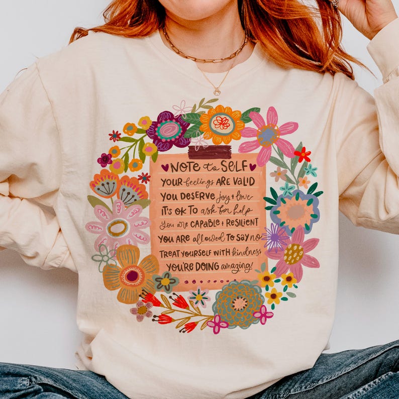 May include: Cream-colored sweatshirt with a floral design and the text "NOTE to SELF." The text includes affirmations such as "Your feelings are valid" and "You deserve joy & love." The design features colorful flowers.