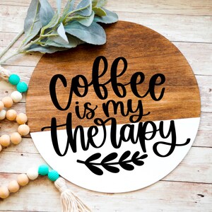 K&ouml;nnte beinhalten: Ein rundes Holzschild mit einem wei&szlig;en Halbkreis, der untenaufgemalt ist. Das Schild sagt "Coffee is my therapy" in schwarzer Schrift mit einem kleinen schwarzen Blattdesign unter dem Text.