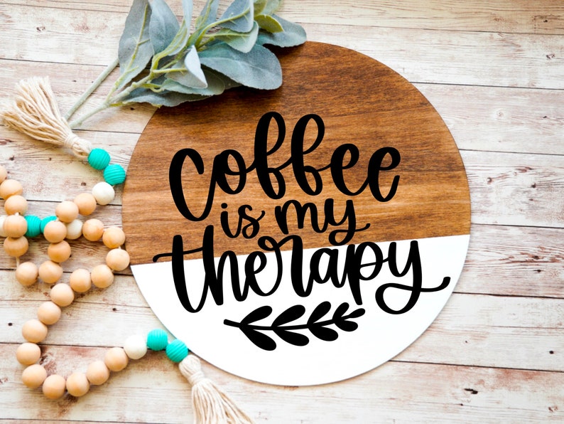 K&ouml;nnte beinhalten: Ein rundes Holzschild mit einem wei&szlig;en Halbkreis, der untenaufgemalt ist. Das Schild sagt "Coffee is my therapy" in schwarzer Schrift mit einem kleinen schwarzen Blattdesign unter dem Text.