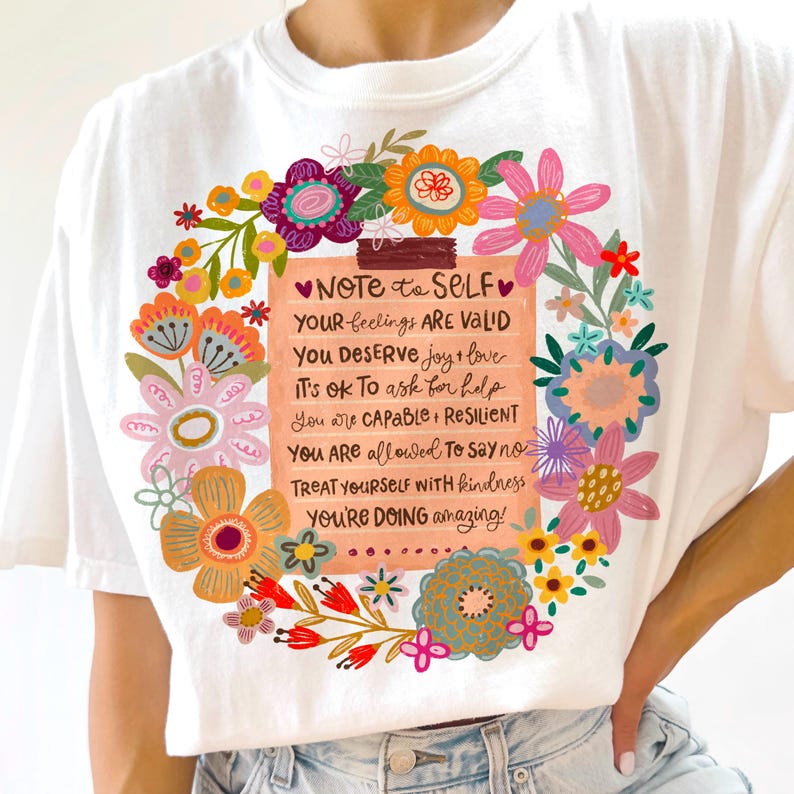 May include: White t-shirt featuring a floral wreath design surrounding a note with uplifting text. The note reads "Note to Self" followed by positive affirmations. The floral wreath includes various colorful flowers.