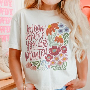 May include: White t-shirt with a floral design and the text "bloom where you are planted".
