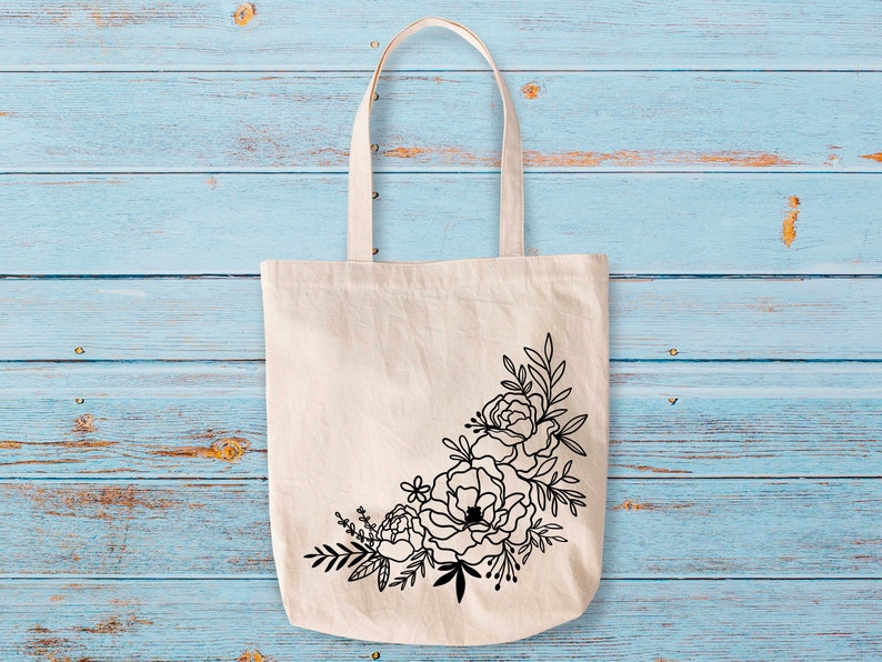 以下が含まれることがあります： 黒い花柄の白いキャンバス地のトートバッグ。デザインはバッグの角に花と葉の集まりが描かれている。