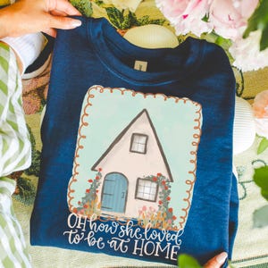 May include: A navy blue sweatshirt featuring a house illustration with a blue door and windows, surrounded by flowers. The text "OH how she loved to be at HOME" is below the house. The sweatshirt is held by a person.