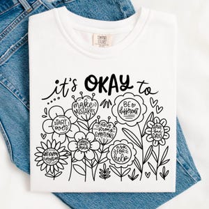 May include: White t-shirt with black text that reads "it's OKAY to" and features a design of flowers with positive affirmations inside each flower. The affirmations include "make mistakes", "be different", "have strong emotions", "start over", "change your mind", "have bad days", "ask for help", and "not know everything".