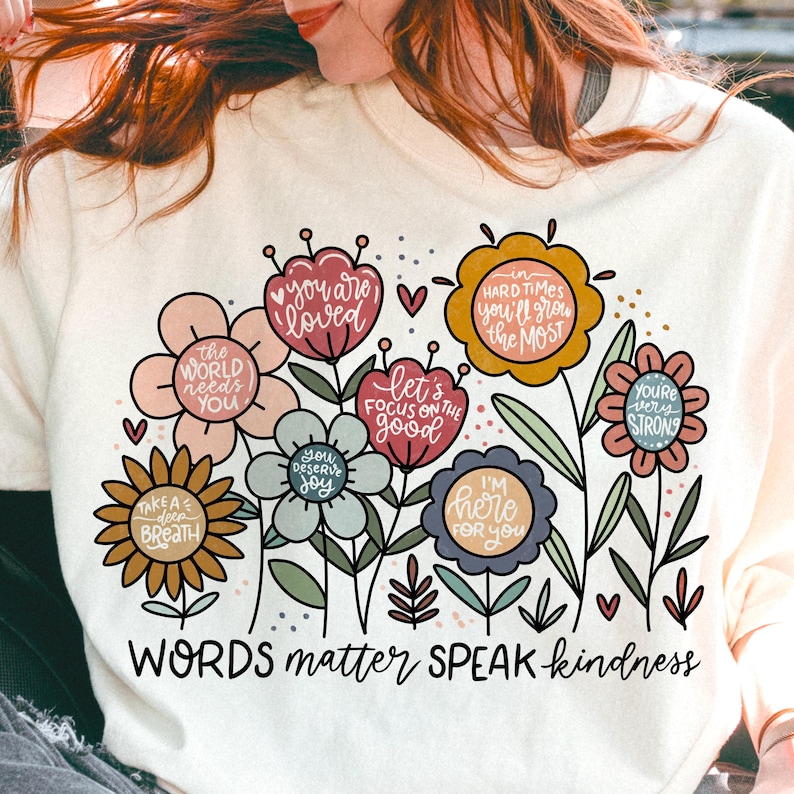 May include: A white t-shirt with a floral design and the text "WORDS matter SPEAK kindness". The flowers are in various colors, including pink, yellow, blue, and green. Each flower has a positive message written inside.