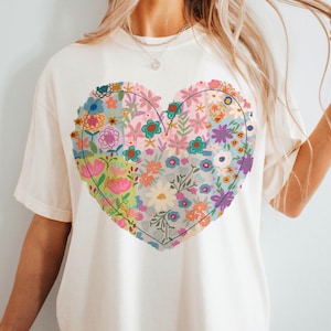 May include: A white t-shirt with a large floral heart design. The heart is made up of different colored flowers and leaves, and is outlined in black.
