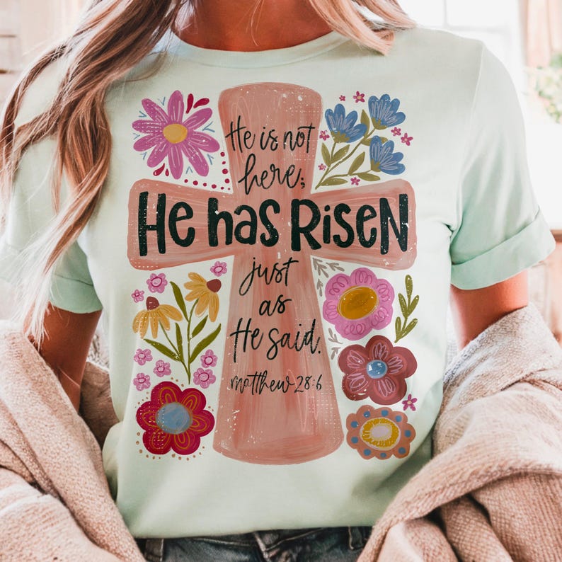 Puede incluir: Camiseta verde claro con un dise&ntilde;o de cruz y las palabras "He has Risen" y "He is not here" en letras negras. La cruz est&aacute; adornada con flores rosas, azules, amarillas y rojas. Tambi&eacute;n se incluye el texto "Matthew 28:6".
