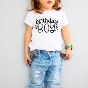 Op de afbeelding: Een wit T-shirt met zwarte tekst die "birthday boy" zegt met een stervormig ontwerp aan beide kanten van het woord "boy".