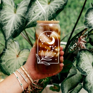 May include: A clear glass tumbler with a bamboo lid and a glass straw, filled with a dark beverage. The tumbler features a white celestial design with a crescent moon, stars, and constellations. The glass is held in front of lush green foliage.