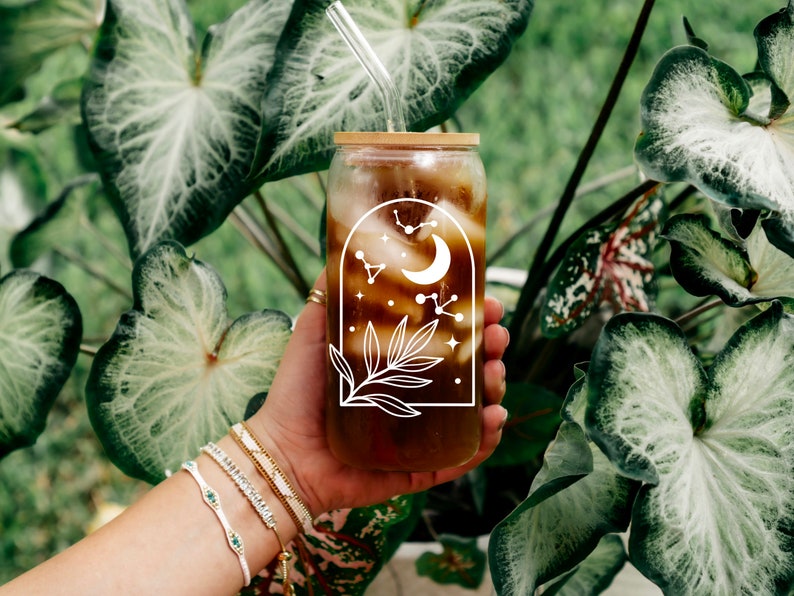 May include: A clear glass tumbler with a bamboo lid and a glass straw, filled with a dark beverage. The tumbler features a white celestial design with a crescent moon, stars, and constellations. The glass is held in front of lush green foliage.