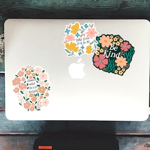 May include: Three floral stickers with inspirational quotes on a white laptop. The stickers feature pink, yellow, and green flowers with black outlines. The quotes are "Take time just to be", "Be Kind", and "Bloom wild, burn bright".
