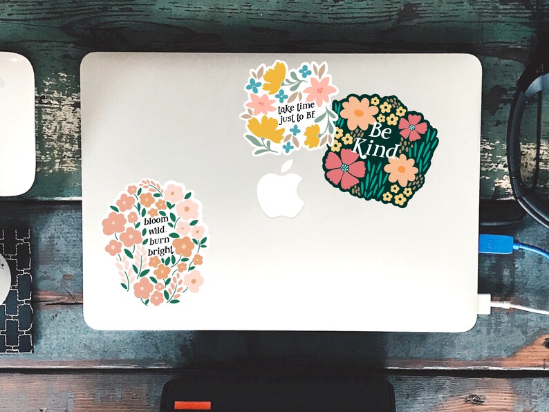May include: Three floral stickers with inspirational quotes on a white laptop. The stickers feature pink, yellow, and green flowers with black outlines. The quotes are "Take time just to be", "Be Kind", and "Bloom wild, burn bright".