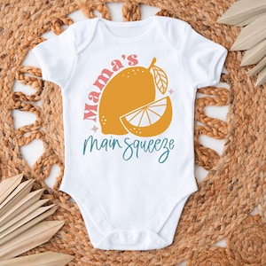 K&ouml;nnte beinhalten: Wei&szlig;er Baby-Body mit einer gelben Zitronengrafik und dem Schriftzug "Mama's Main Squeeze" in Rosa und T&uuml;rkis. Der Body wird auf einer gewebten Matte mit getrockneten Palmbl&auml;ttern pr&auml;sentiert.