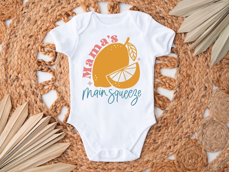 K&ouml;nnte beinhalten: Wei&szlig;er Baby-Body mit einer gelben Zitronengrafik und dem Schriftzug "Mama's Main Squeeze" in Rosa und T&uuml;rkis. Der Body wird auf einer gewebten Matte mit getrockneten Palmbl&auml;ttern pr&auml;sentiert.