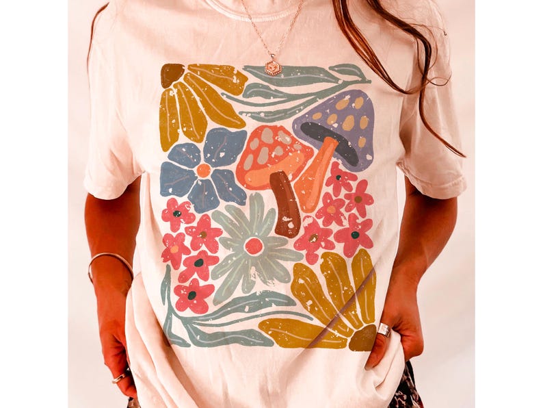 May include: A white t-shirt with a colorful floral and mushroom graphic print. The design features a variety of flowers, leaves, and mushrooms in shades of red, blue, yellow, and green.