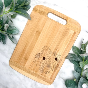 May include: A natural bamboo cutting board with a floral design engraved on the surface. The board has a handle for easy carrying.