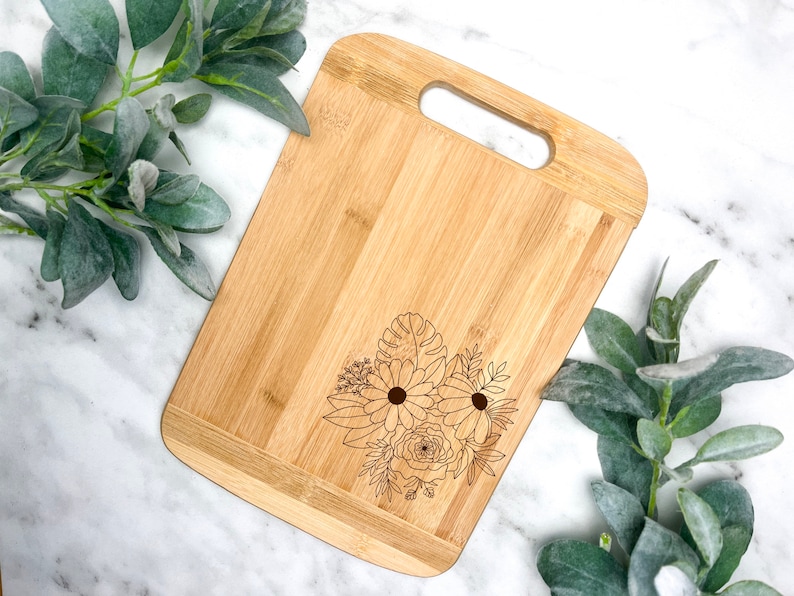 May include: A natural bamboo cutting board with a floral design engraved on the surface. The board has a handle for easy carrying.