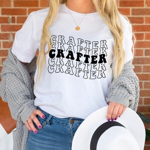 May include: White t-shirt with the word "CRAFTER" repeated in a retro font style. The shirt is worn by a person with long blonde hair and a gray cardigan.