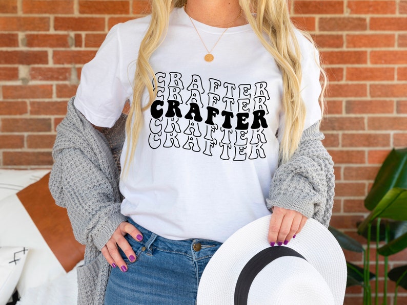 May include: White t-shirt with the word "CRAFTER" repeated in a retro font style. The shirt is worn by a person with long blonde hair and a gray cardigan.