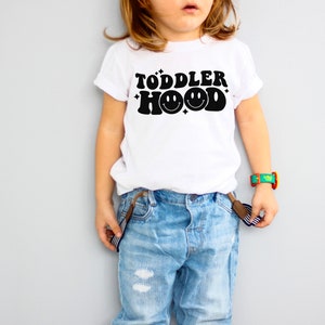 K&ouml;nnte beinhalten: Wei&szlig;es T-Shirt mit dem schwarzen Text "TODDLER HOOD" und einem Smiley-Gesicht. Das Kind tr&auml;gt eine blaue Jeans und Hosentr&auml;ger. Eine bunte Uhr befindet sich am Handgelenk des Kindes.
