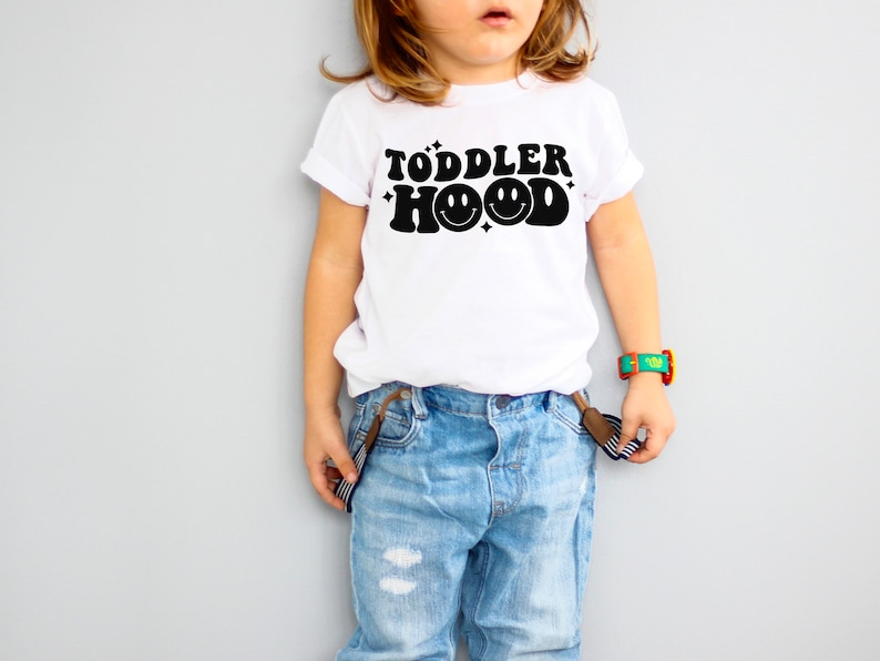 K&ouml;nnte beinhalten: Wei&szlig;es T-Shirt mit dem schwarzen Text "TODDLER HOOD" und einem Smiley-Gesicht. Das Kind tr&auml;gt eine blaue Jeans und Hosentr&auml;ger. Eine bunte Uhr befindet sich am Handgelenk des Kindes.