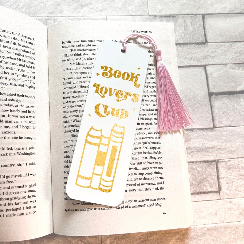 Pode incluir: Marcador de livros branco com texto dourado "Book Lovers Club" e um gr&aacute;fico de livros. Uma borla rosa pende do topo. O marcador de livros est&aacute; colocado num livro aberto.