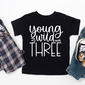 Op de afbeelding: Zwart T-shirt met witte tekst die "young wild and three" luidt. Het shirt ligt op een witte ondergrond met een geruit overhemd, een blauwe spijkerbroek en zwarte sneakers.