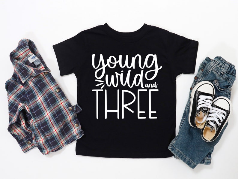 Op de afbeelding: Zwart T-shirt met witte tekst die "young wild and three" luidt. Het shirt ligt op een witte ondergrond met een geruit overhemd, een blauwe spijkerbroek en zwarte sneakers.