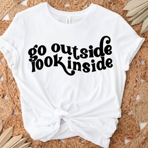 May include: White t-shirt with black text that reads "go outside look inside" in a retro font.