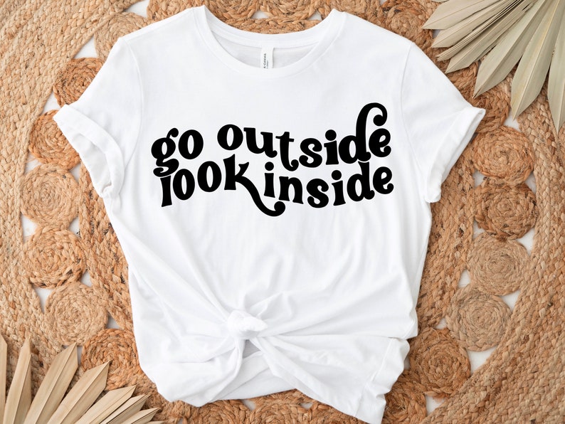 May include: White t-shirt with black text that reads "go outside look inside" in a retro font.