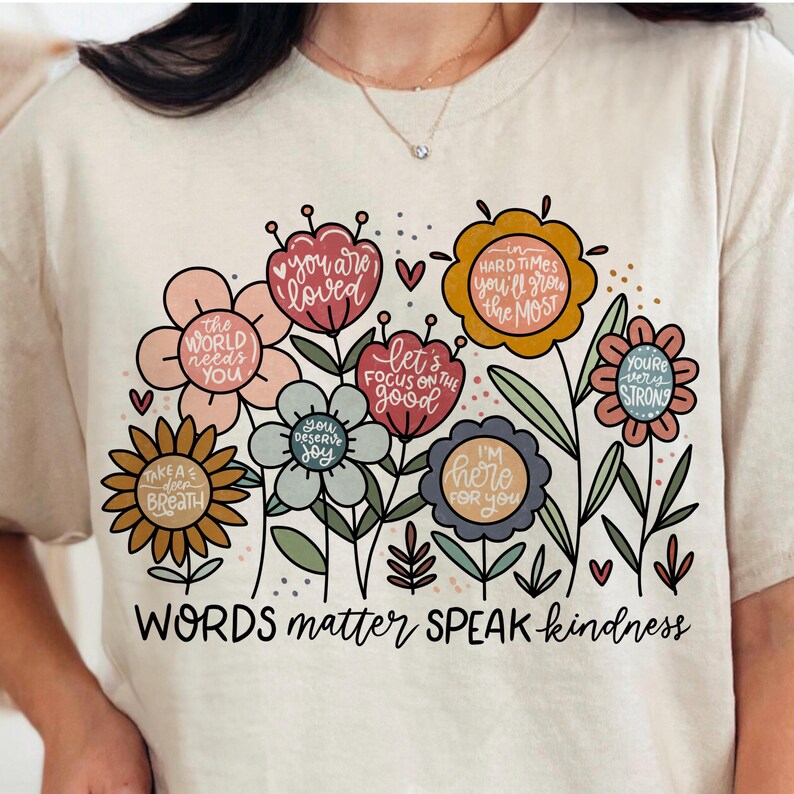May include: A beige t-shirt with a floral design and the text "WORDS matter SPEAK kindness". The flowers are in various colors, including pink, yellow, blue, and green. Each flower has a different inspirational message inside.