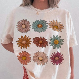 May include: White t-shirt with a floral print featuring nine colorful daisies in a grid pattern.