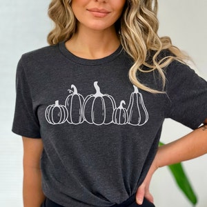 May include: A dark grey t-shirt featuring a white outline of five pumpkins of varying sizes. The pumpkins are arranged in a row, from largest to smallest.