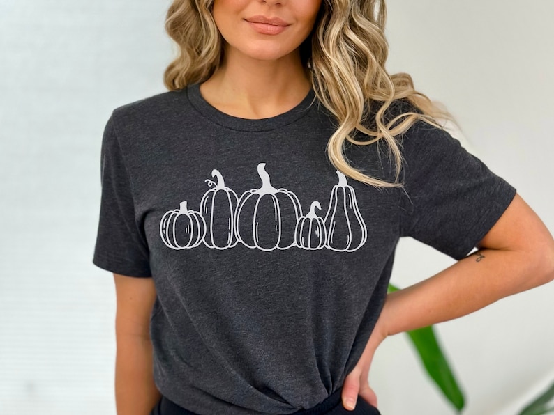 May include: A dark grey t-shirt featuring a white outline of five pumpkins of varying sizes. The pumpkins are arranged in a row, from largest to smallest.