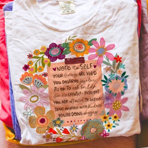 May include: White t-shirt with a floral design surrounding a note that reads "Note to Self." The note includes positive affirmations such as "Your feelings are valid" and "You're doing amazing!"