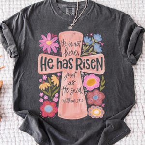 Puede incluir: Camiseta gris oscuro con un dise&ntilde;o de cruz rosa y las palabras "Ha resucitado" y "No est&aacute; aqu&iacute;." La cruz est&aacute; adornada con flores coloridas. Tambi&eacute;n es visible el texto "Mateo 28:6".