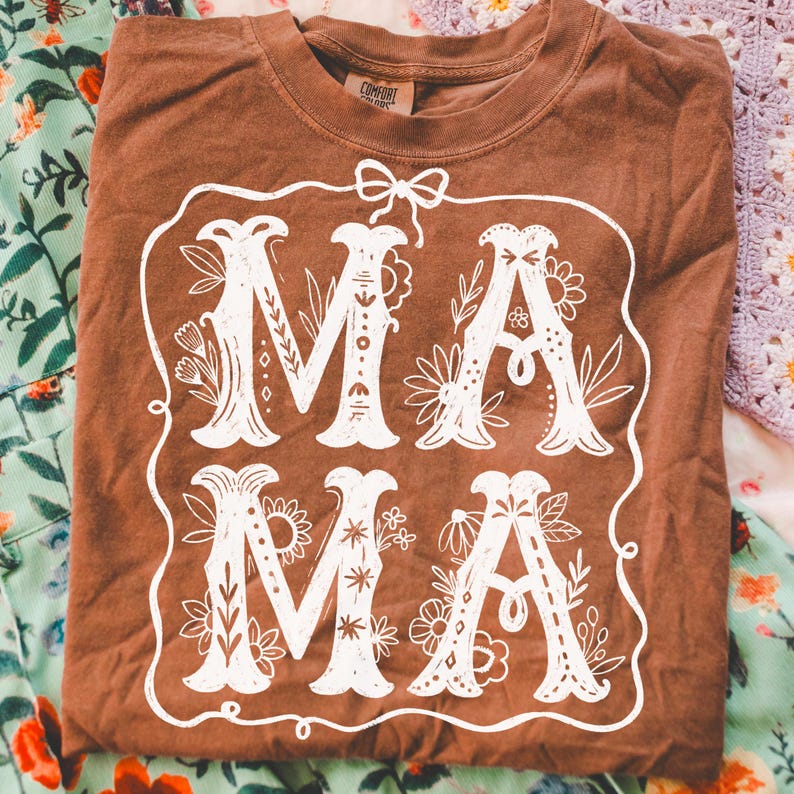 May include: A brown t-shirt with a white floral design that says "MAMA" in a decorative font.