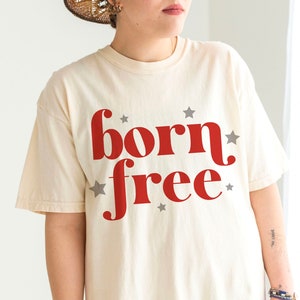 K&ouml;nnte beinhalten: Ein cremefarbenes T-Shirt mit dem Aufdruck "born free" in Rot. Die W&ouml;rter sind in einer Retro-Schriftart gehalten und von kleinen grauen Sternen umgeben.