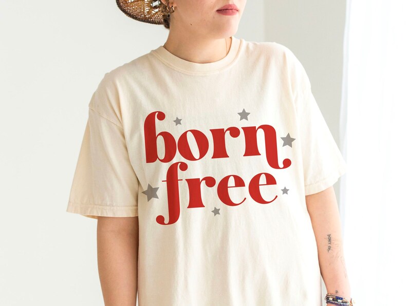 K&ouml;nnte beinhalten: Ein cremefarbenes T-Shirt mit dem Aufdruck "born free" in Rot. Die W&ouml;rter sind in einer Retro-Schriftart gehalten und von kleinen grauen Sternen umgeben.