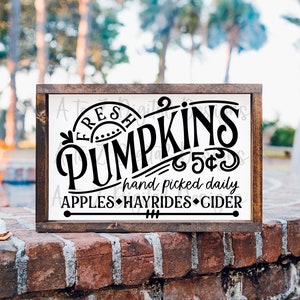 May include: A white wooden sign with black lettering that reads "Fresh Pumpkins 5&cent; hand picked daily Apples Hayrides Cider". The sign is framed in dark brown wood and is sitting on a brick wall.
