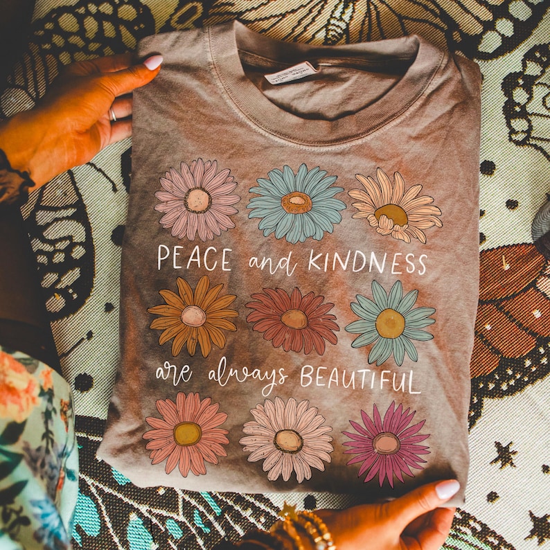 K&ouml;nnte beinhalten: Ein braunes T-Shirt mit einem Batik-Effekt zeigt ein Design mit bunten Blumen und dem Text "Peace and kindness are always beautiful."