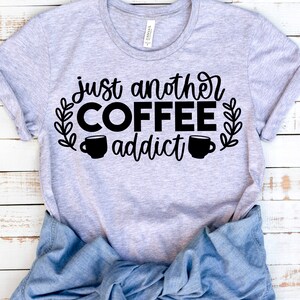 K&ouml;nnte beinhalten: Ein graues T-Shirt mit dem Text "just another coffee addict" in schwarzer Schrift. Der Text ist von zwei Kaffeetassen-Symbolen und Blattwerk umgeben.