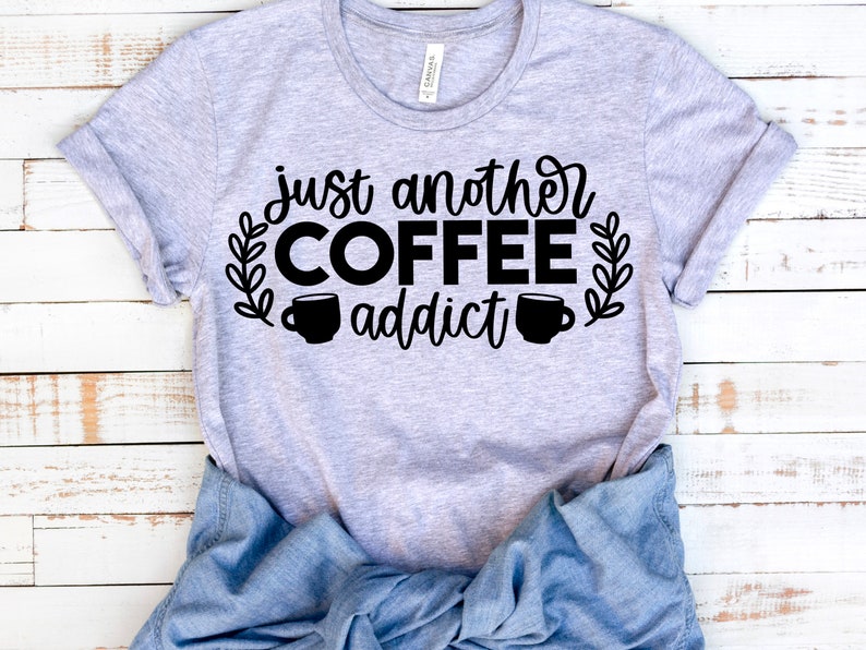 K&ouml;nnte beinhalten: Ein graues T-Shirt mit dem Text "just another coffee addict" in schwarzer Schrift. Der Text ist von zwei Kaffeetassen-Symbolen und Blattwerk umgeben.