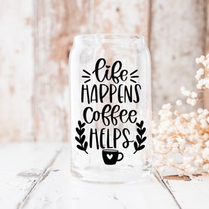 K&ouml;nnte beinhalten: Ein transparentes Glasgef&auml;&szlig; mit einer schwarzen Grafik, auf der "life happens coffee helps" steht, sowie einem kleinen Kaffeetassen-Symbol. Das Gef&auml;&szlig; steht auf einer wei&szlig;en Holzoberfl&auml;che.