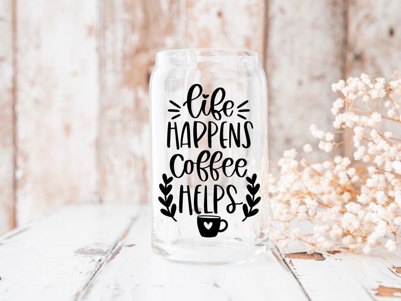 K&ouml;nnte beinhalten: Ein transparentes Glasgef&auml;&szlig; mit einer schwarzen Grafik, auf der "life happens coffee helps" steht, sowie einem kleinen Kaffeetassen-Symbol. Das Gef&auml;&szlig; steht auf einer wei&szlig;en Holzoberfl&auml;che.