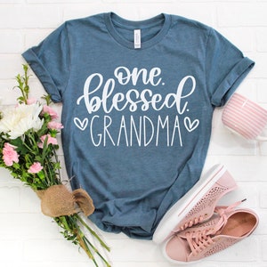 May include: A blue t-shirt with the text "one blessed grandma" in white lettering with two white hearts on either side of the word "grandma".